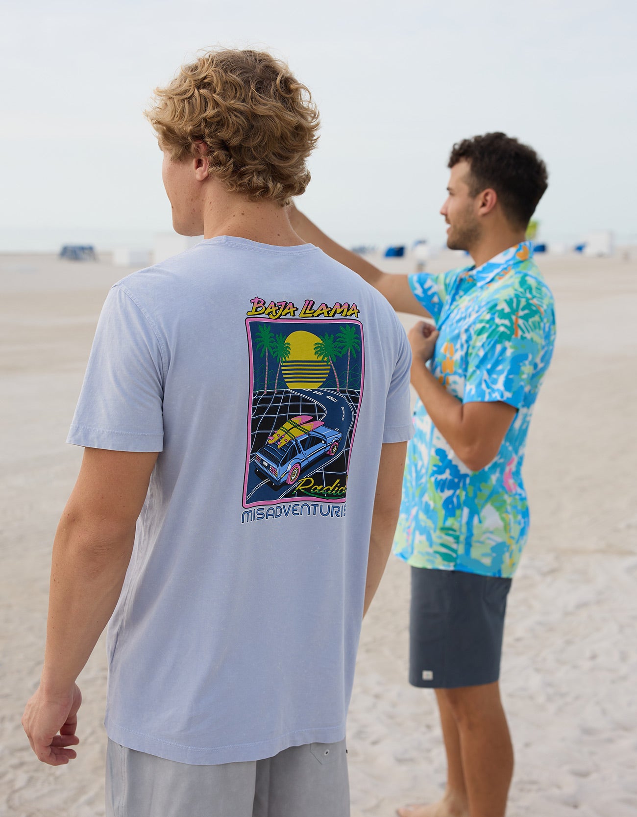 Model wearing Baja Llama Radical Misadventurist purple acid wash graphic tee showing back print on the beach with another model in background