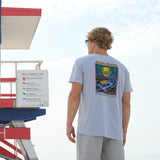 Model standing near a beach warning sign wearing the Radical Misadventurist purple acid wash graphic tee showing back print
