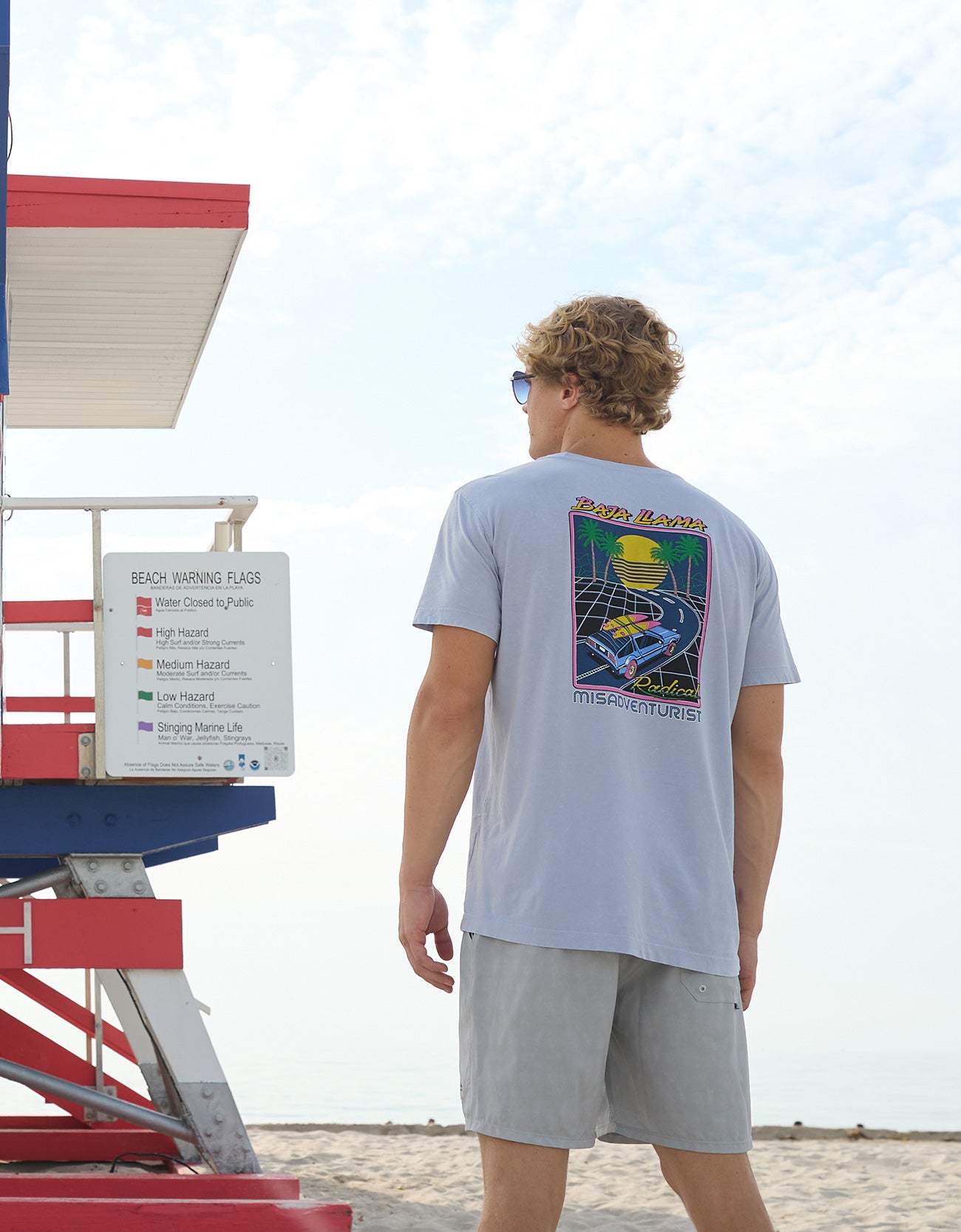Model standing near a beach warning sign wearing the Radical Misadventurist purple acid wash graphic tee showing back print