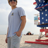 Model wearing the Radical Misadventurist purple acid wash graphic tee and sunglasses on the sand next to an American flag lifeguard tower