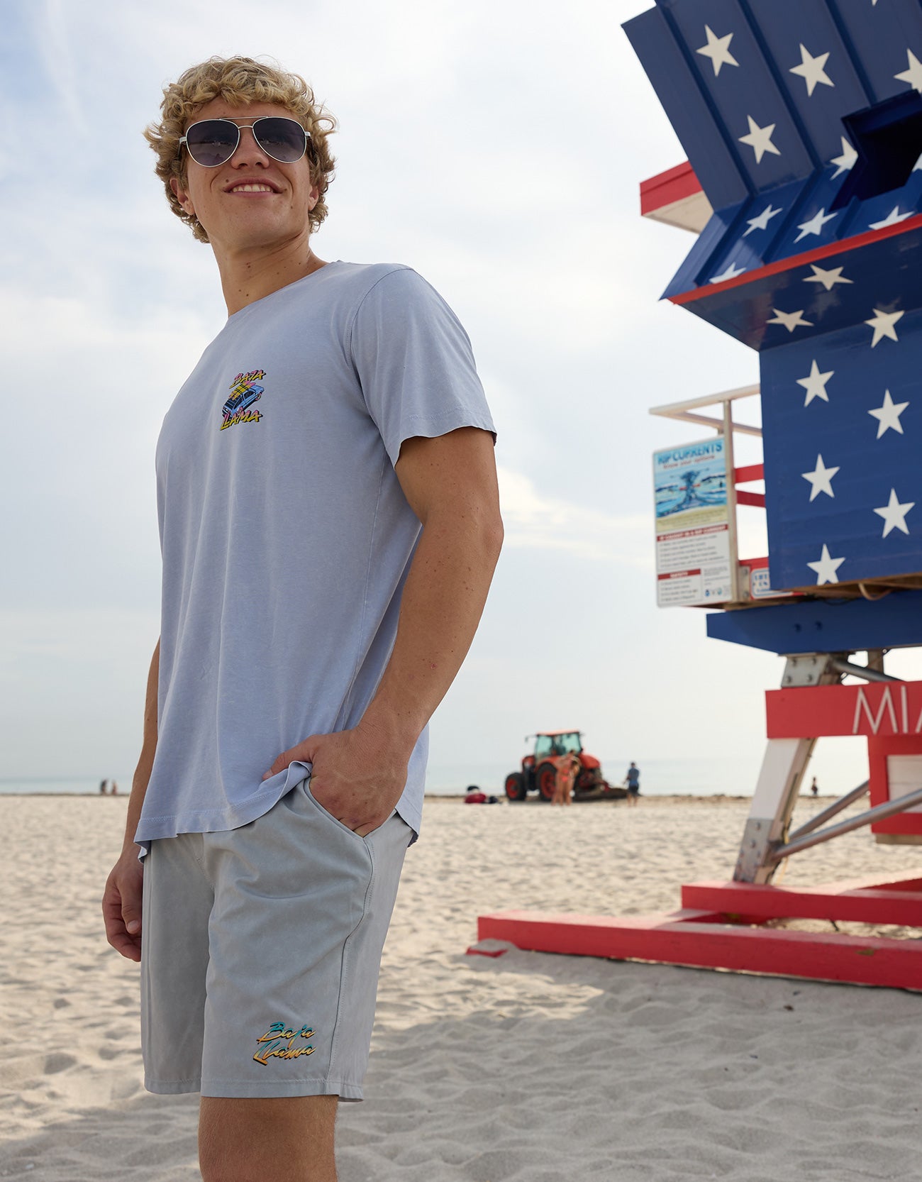 Model wearing the Radical Misadventurist purple acid wash graphic tee and sunglasses on the sand next to an American flag lifeguard tower