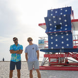 Two models standing on the sand in front of a Miami Beach lifeguard tower wearing Baja Llama apparel including the Radical Misadventurist purple acid wash graphic tee