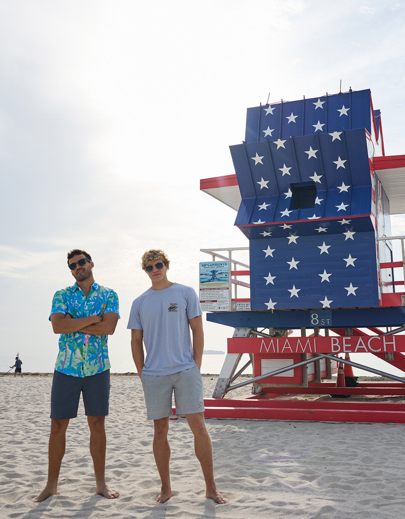 Two models standing on the sand in front of a Miami Beach lifeguard tower wearing Baja Llama apparel including the Radical Misadventurist purple acid wash graphic tee