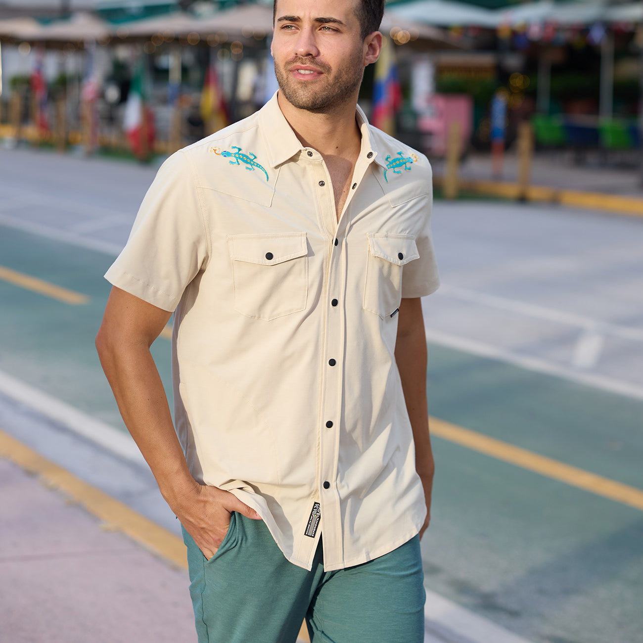 Model walking outdoors wearing the Baja Llama Reptile Wrangler cream western snap button-up shirt with embroidered gecko shoulder details and teal shorts