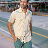 Model walking outdoors wearing the Baja Llama Reptile Wrangler cream western snap button-up shirt with embroidered gecko shoulder details and teal shorts
