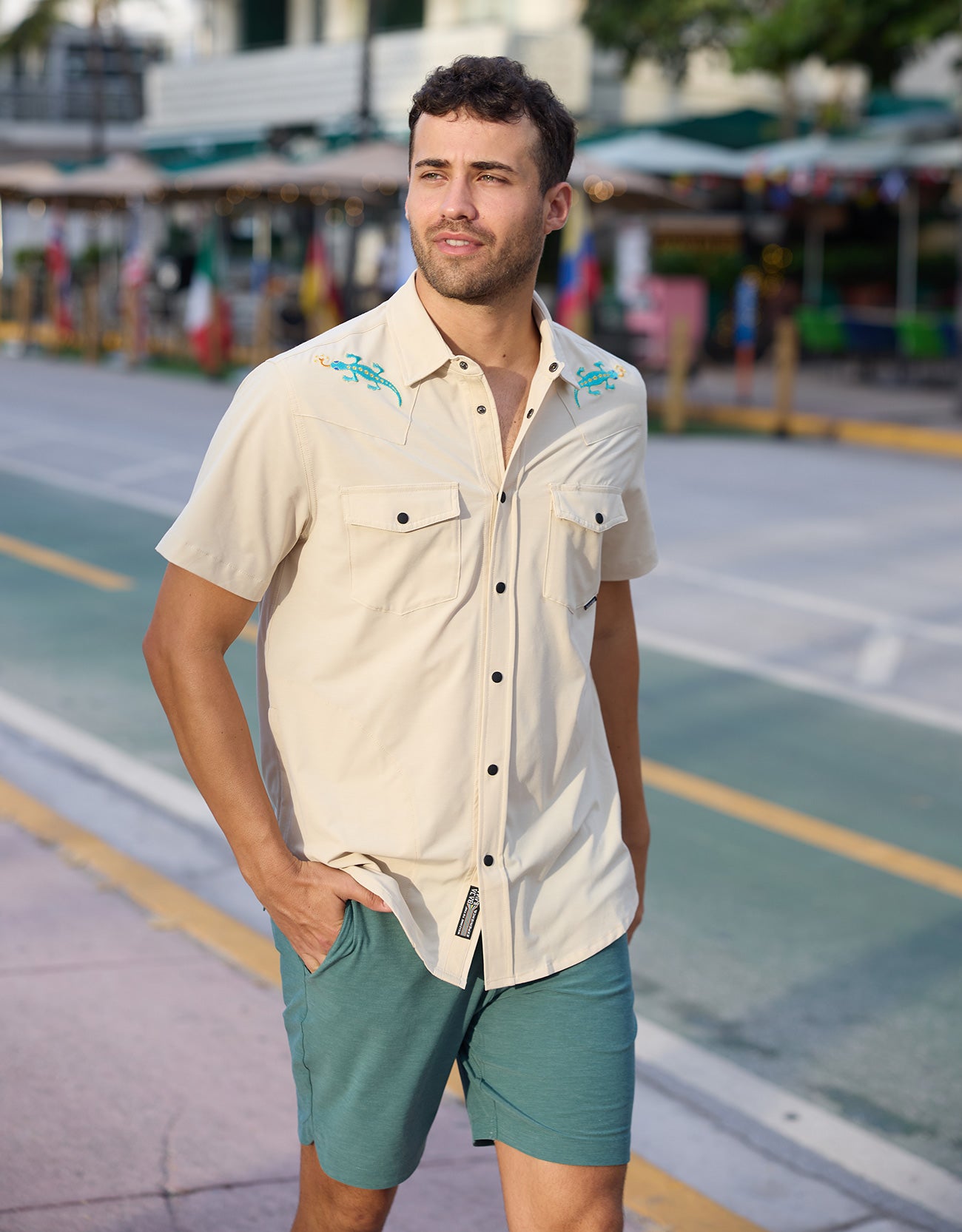 Model walking outdoors wearing the Baja Llama Reptile Wrangler cream western snap button-up shirt with embroidered gecko shoulder details and teal shorts