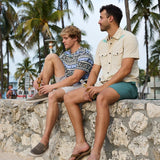 Two models sitting on a stone wall under palm trees holding bottled drinks, wearing Baja Llama apparel including the Reptile Wrangler cream western button-up shirt