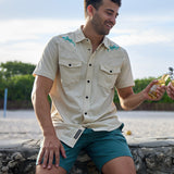 Smiling model sitting outdoors near the beach holding a bottled drink wearing the Baja Llama Reptile Wrangler cream western button-up shirt with embroidered gecko shoulder details