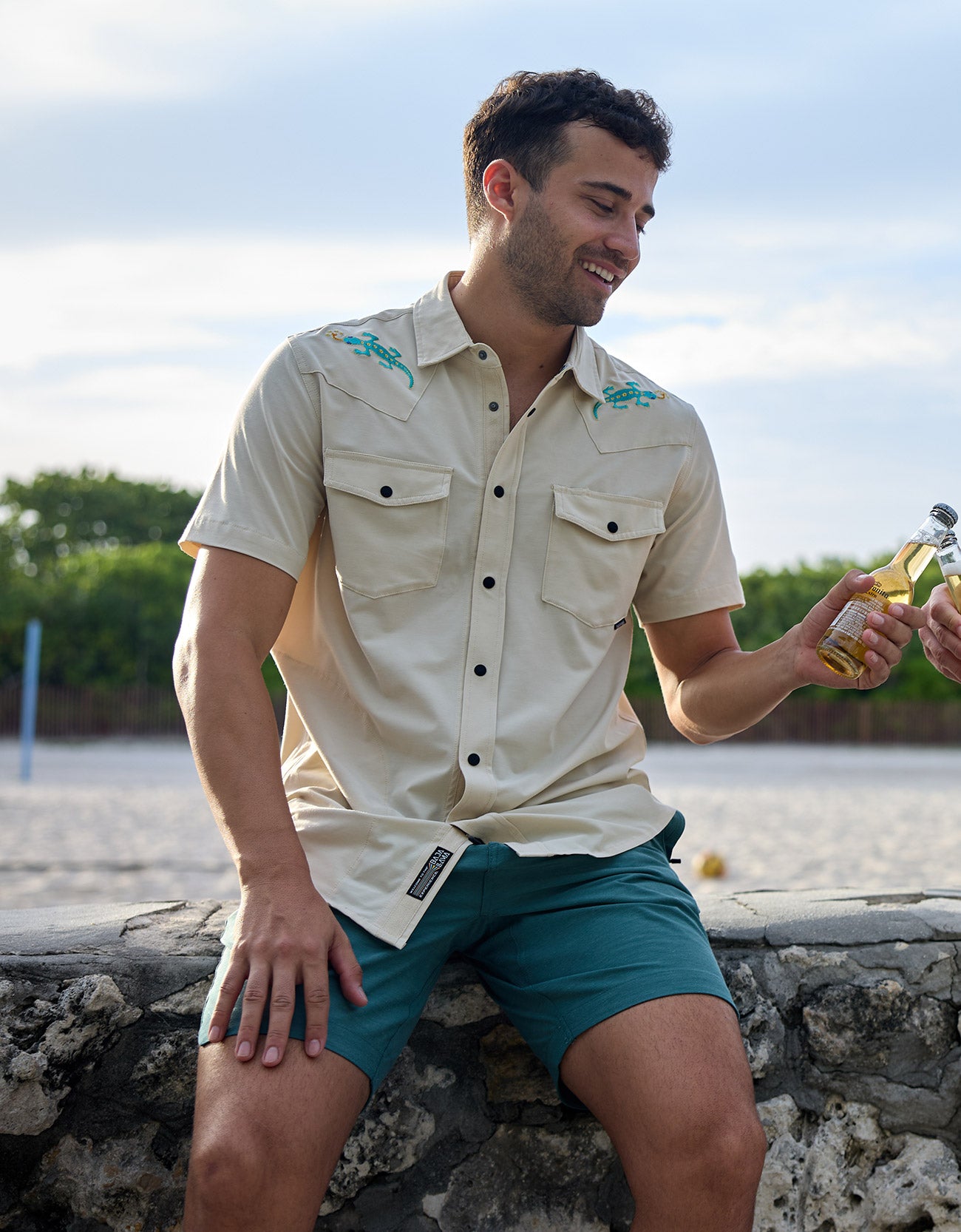Smiling model sitting outdoors near the beach holding a bottled drink wearing the Baja Llama Reptile Wrangler cream western button-up shirt with embroidered gecko shoulder details