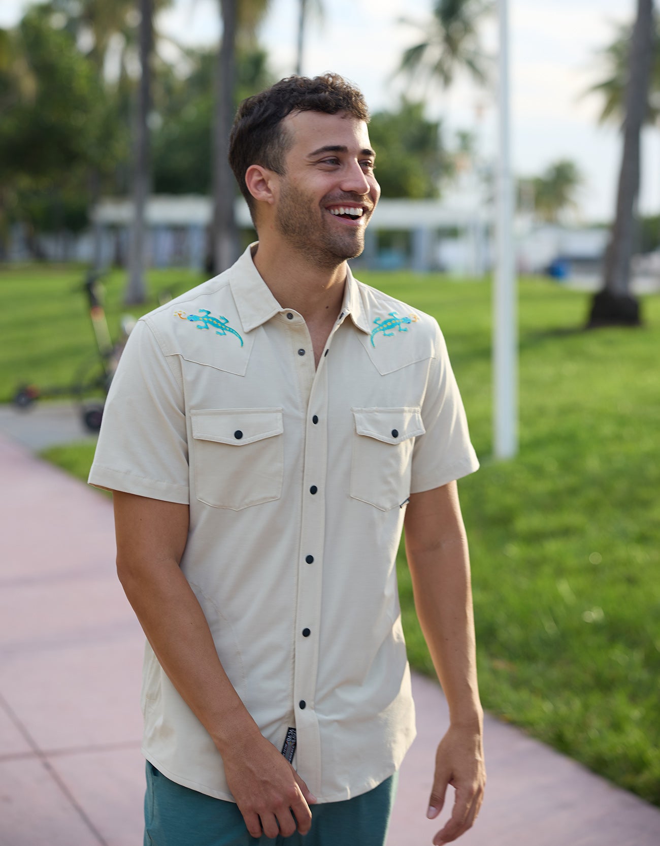 Smiling model walking in a park wearing the Baja Llama Reptile Wrangler cream western button-up shirt with embroidered gecko shoulder details