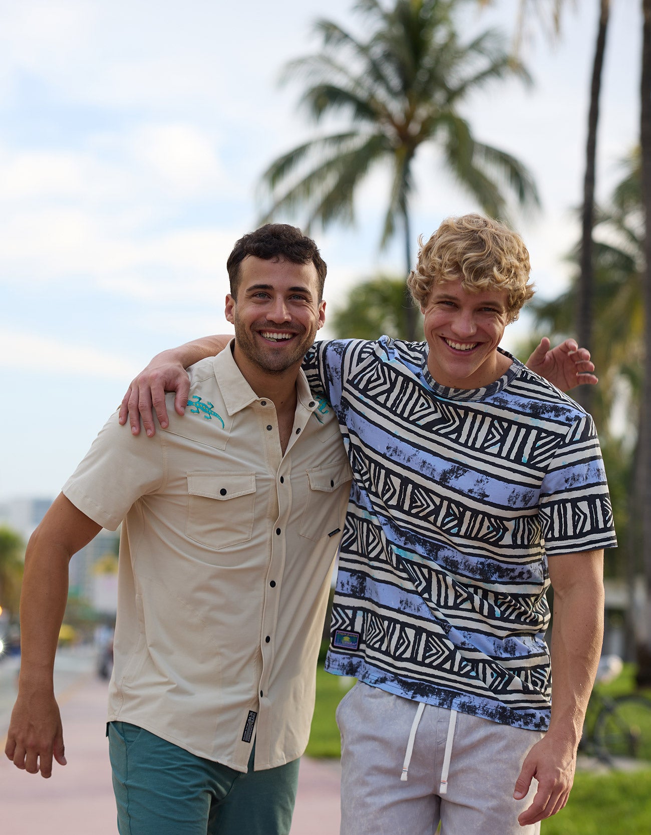 Two smiling male models walking with arms around each other outdoors wearing Baja Llama apparel including the Reptile Wrangler cream western button-up shirt with embroidered gecko accents