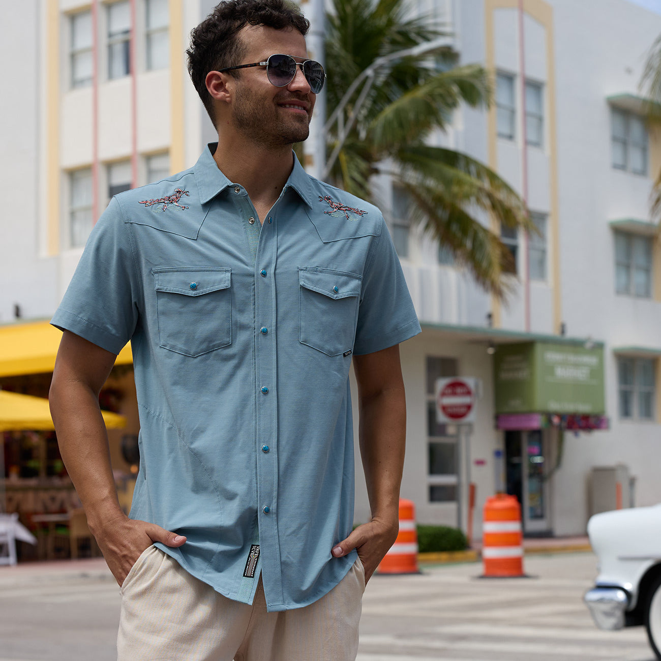 Model wearing the Baja Llama Road Runner blue western button-up shirt with embroidered shoulder details and sunglasses on a city street