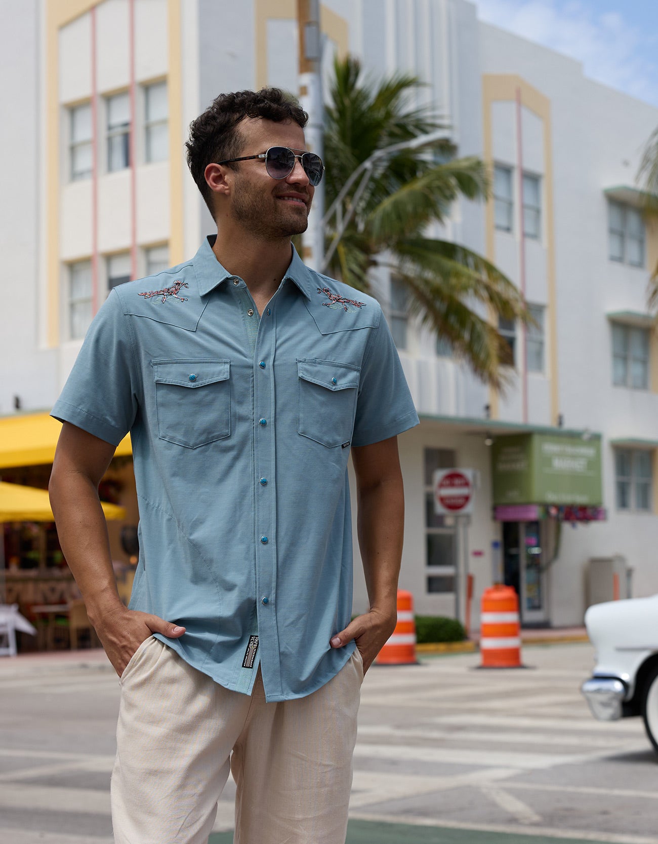 Model wearing the Baja Llama Road Runner blue western button-up shirt with embroidered shoulder details and sunglasses on a city street
