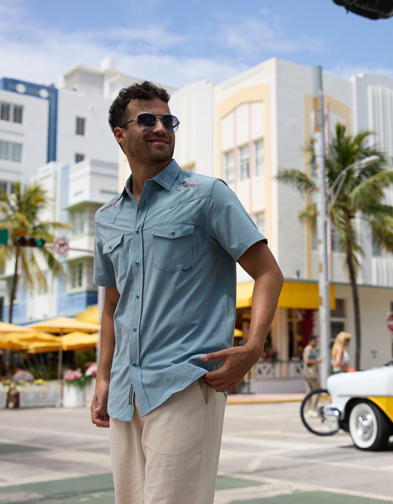 Model wearing the Baja Llama Road Runner blue western snap button-up shirt with embroidered shoulder details and light trousers on a city street