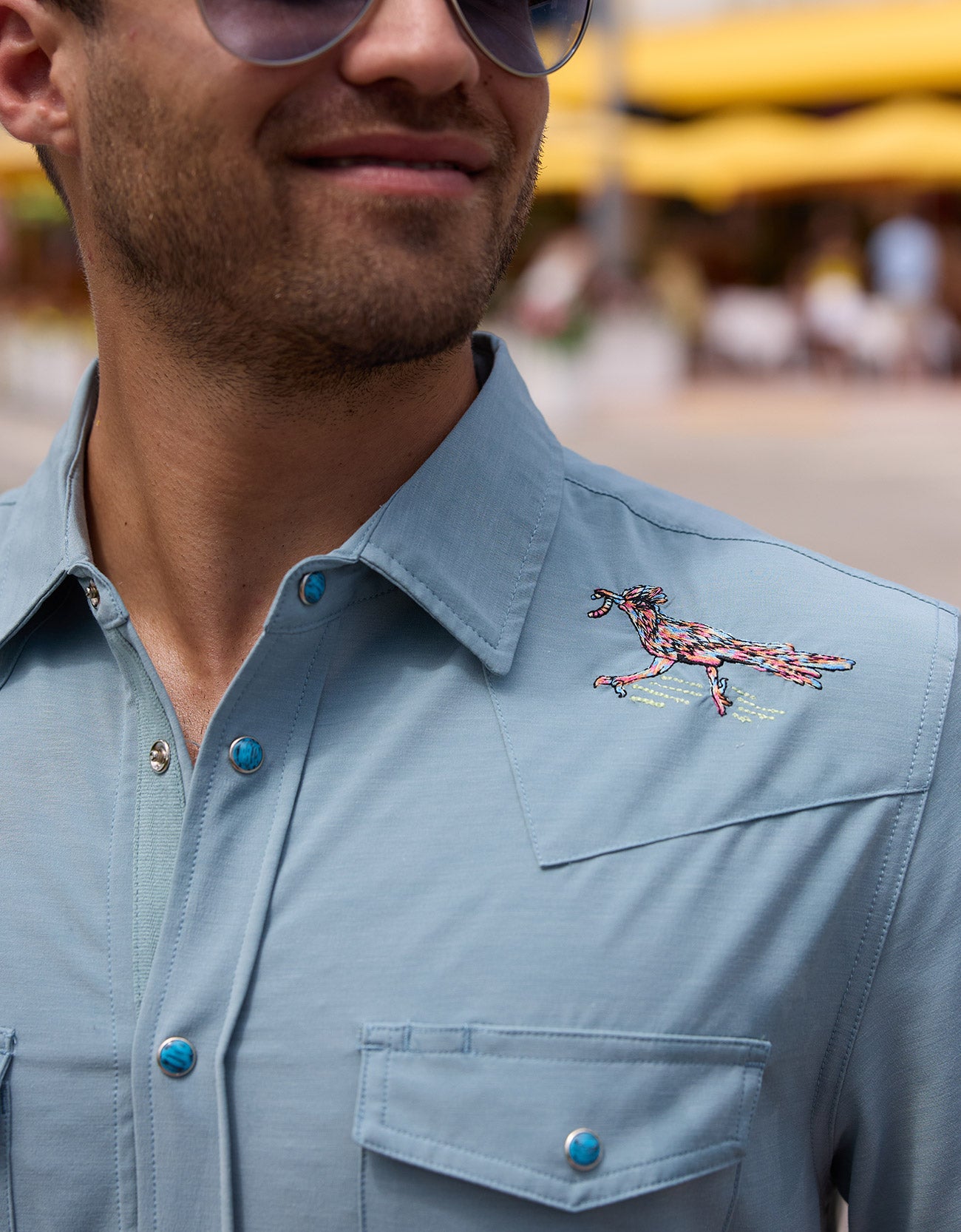 Close-up of the Baja Llama Road Runner blue western snap button-up shirt showing detailed road runner shoulder embroidery and turquoise-accented buttons