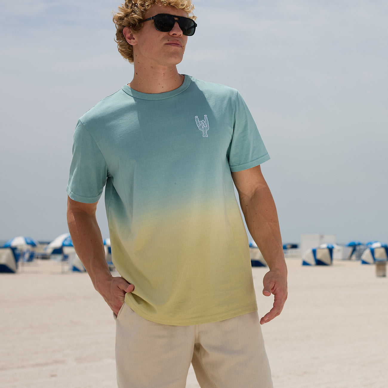 Model standing on a sunny beach wearing sunglasses and the Baja Llama Rock On Cactus embroidered ombre heavy tee in teal and yellow
