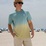 Model standing on a sunny beach wearing sunglasses and the Baja Llama Rock On Cactus embroidered ombre heavy tee in teal and yellow