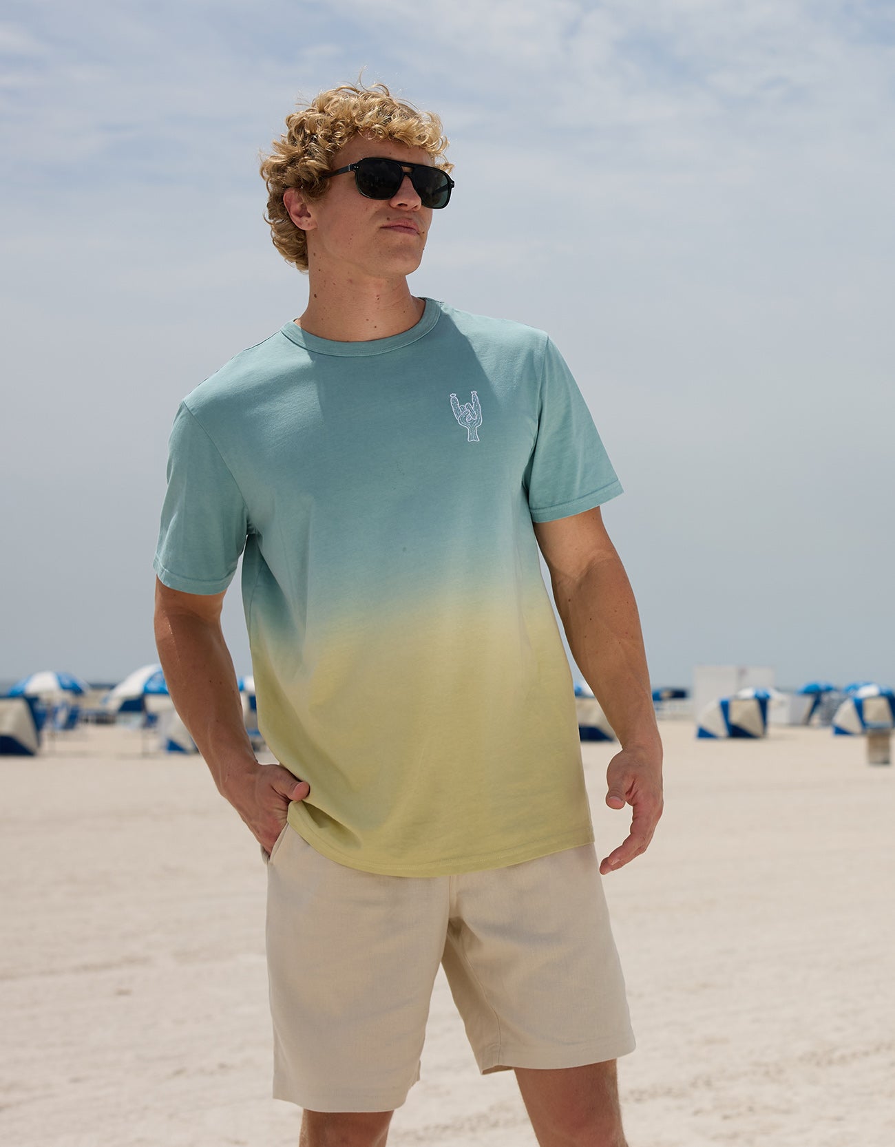 Model standing on a sunny beach wearing sunglasses and the Baja Llama Rock On Cactus embroidered ombre heavy tee in teal and yellow