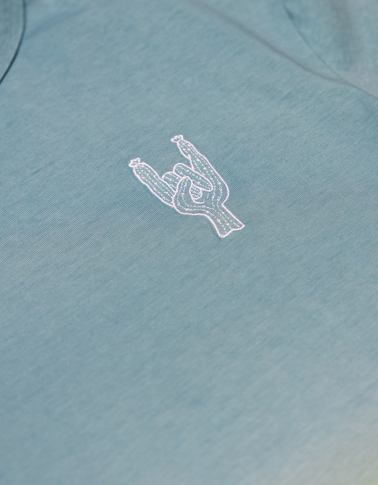 Close-up of the left chest on the Baja Llama Rock On Cactus Embroidered Primo S.E. Heavy Tee, showcasing the small white embroidered rock-on sign cactus against the teal-blue upper section of the shirt.