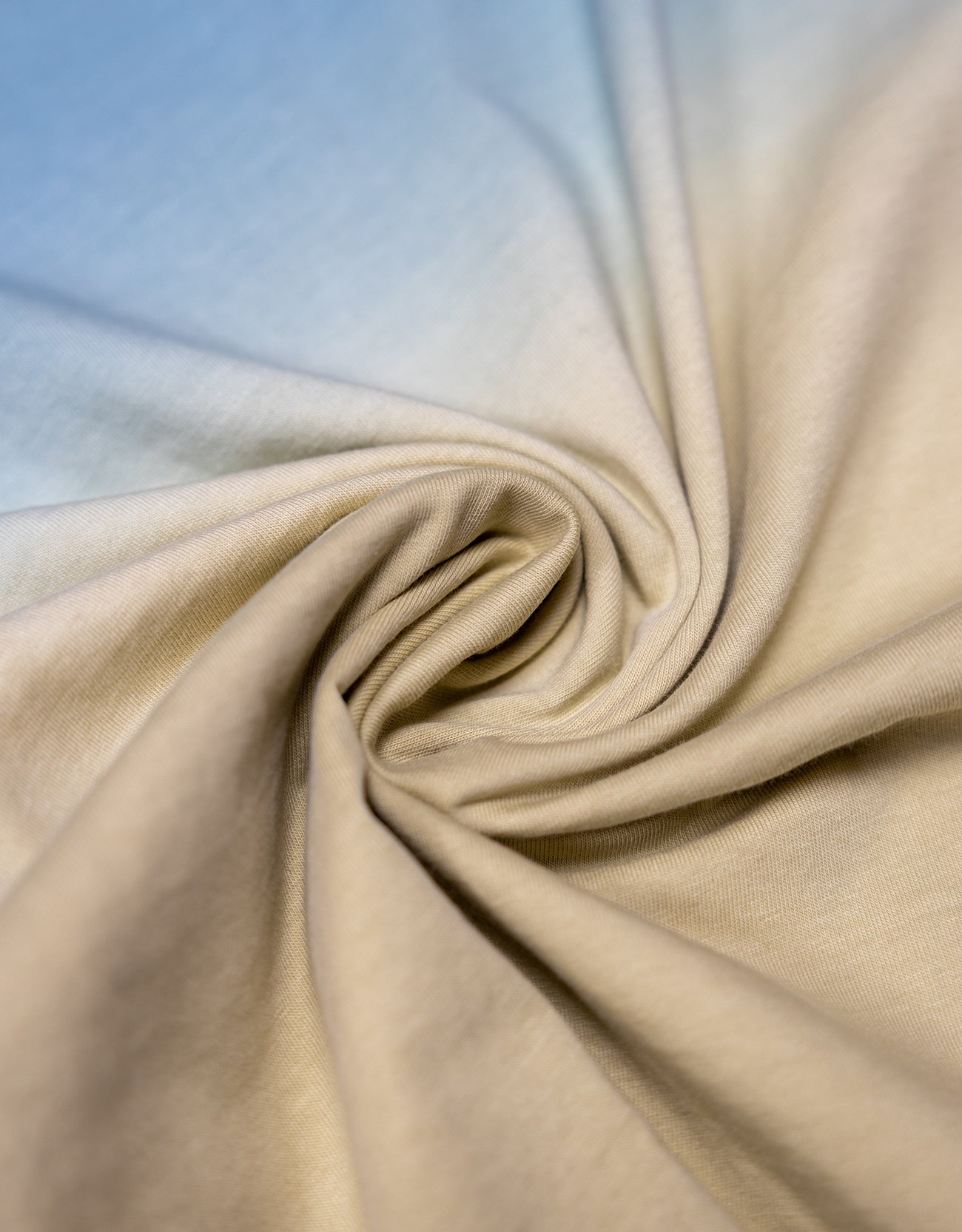 A close-up view of the Baja Llama Rock On Cactus Embroidered Primo S.E. Heavy Tee fabric, tightly swirled to showcase the heavy cotton texture and the smooth gradient transition from ocean blue to sandy tan