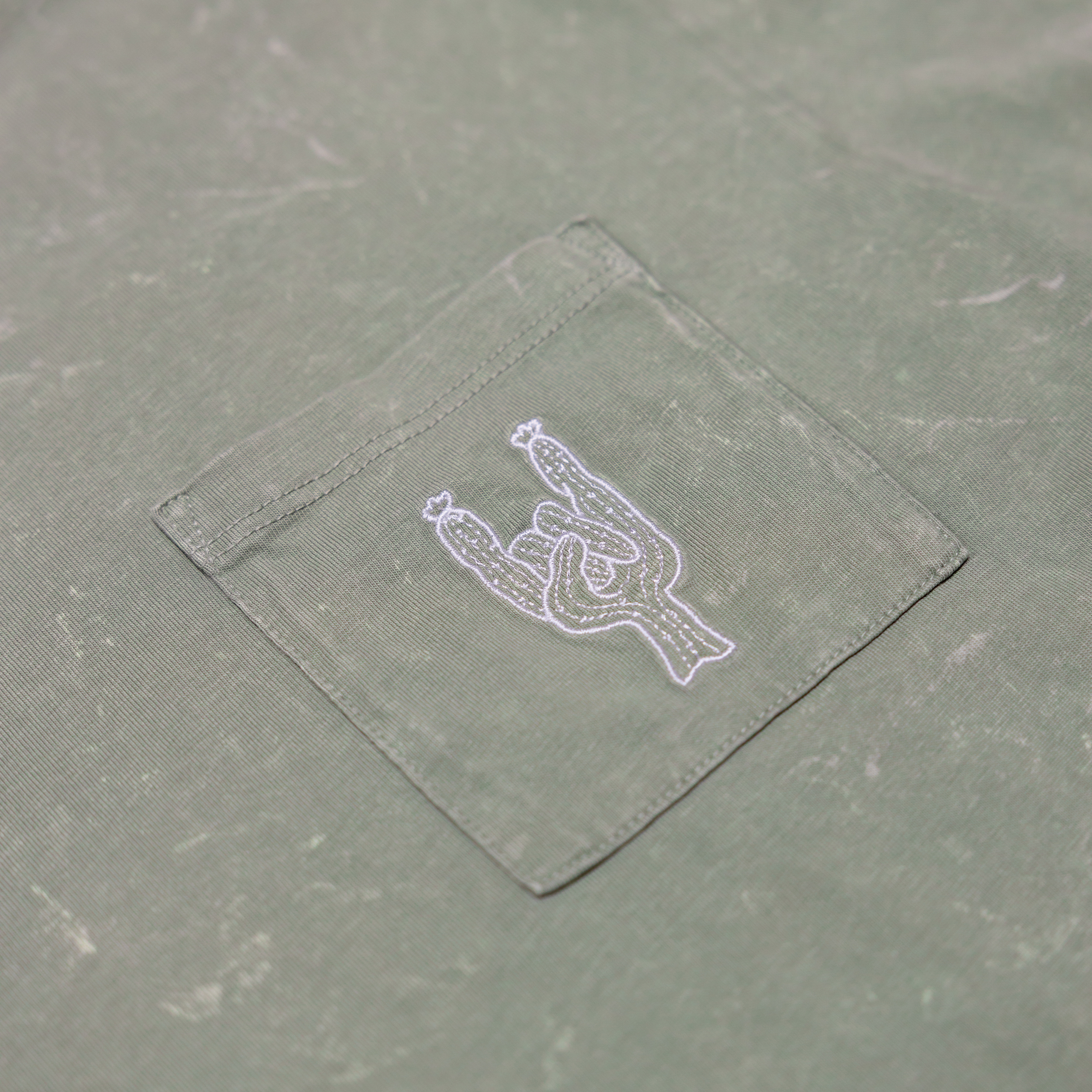 Close-up of the front chest pocket on the Baja Llama Greatful Dyed Rock On Cypress Green Heavy Tee, showing a white embroidered cactus in the shape of a rock-on sign