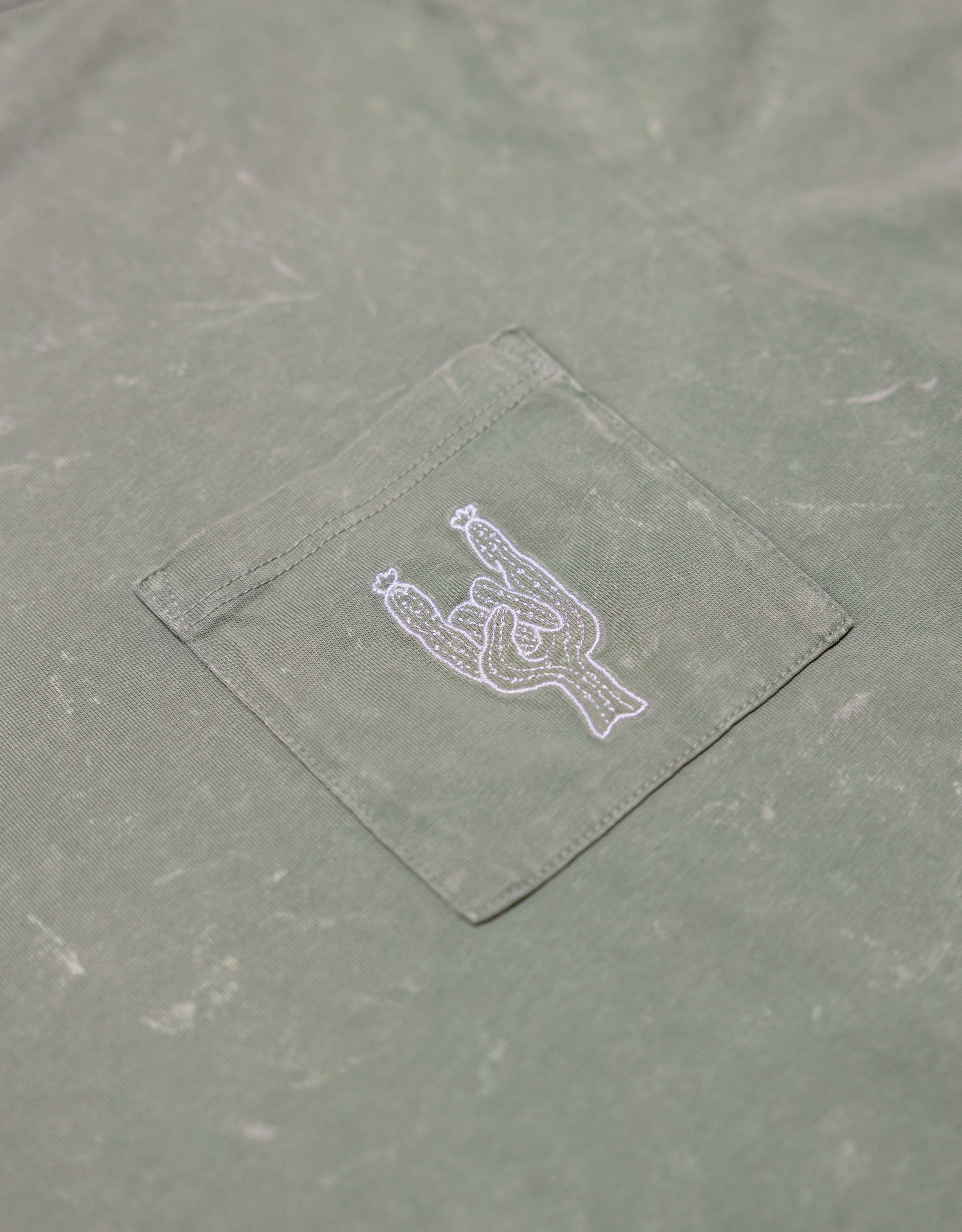 Close-up of the front chest pocket on the Baja Llama Greatful Dyed Rock On Cypress Green Heavy Tee, showing a white embroidered cactus in the shape of a rock-on sign