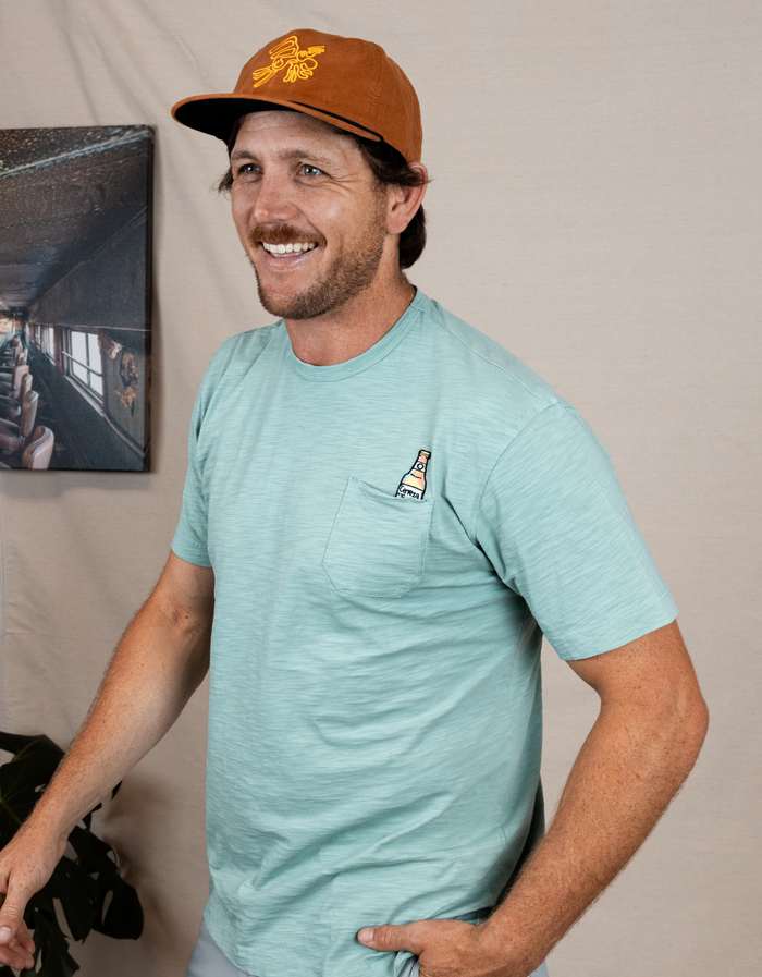 Lifestyle smiling view of mint Cerveza Slub Pima pocket tee with cerveza bottle embroidery and rust hat