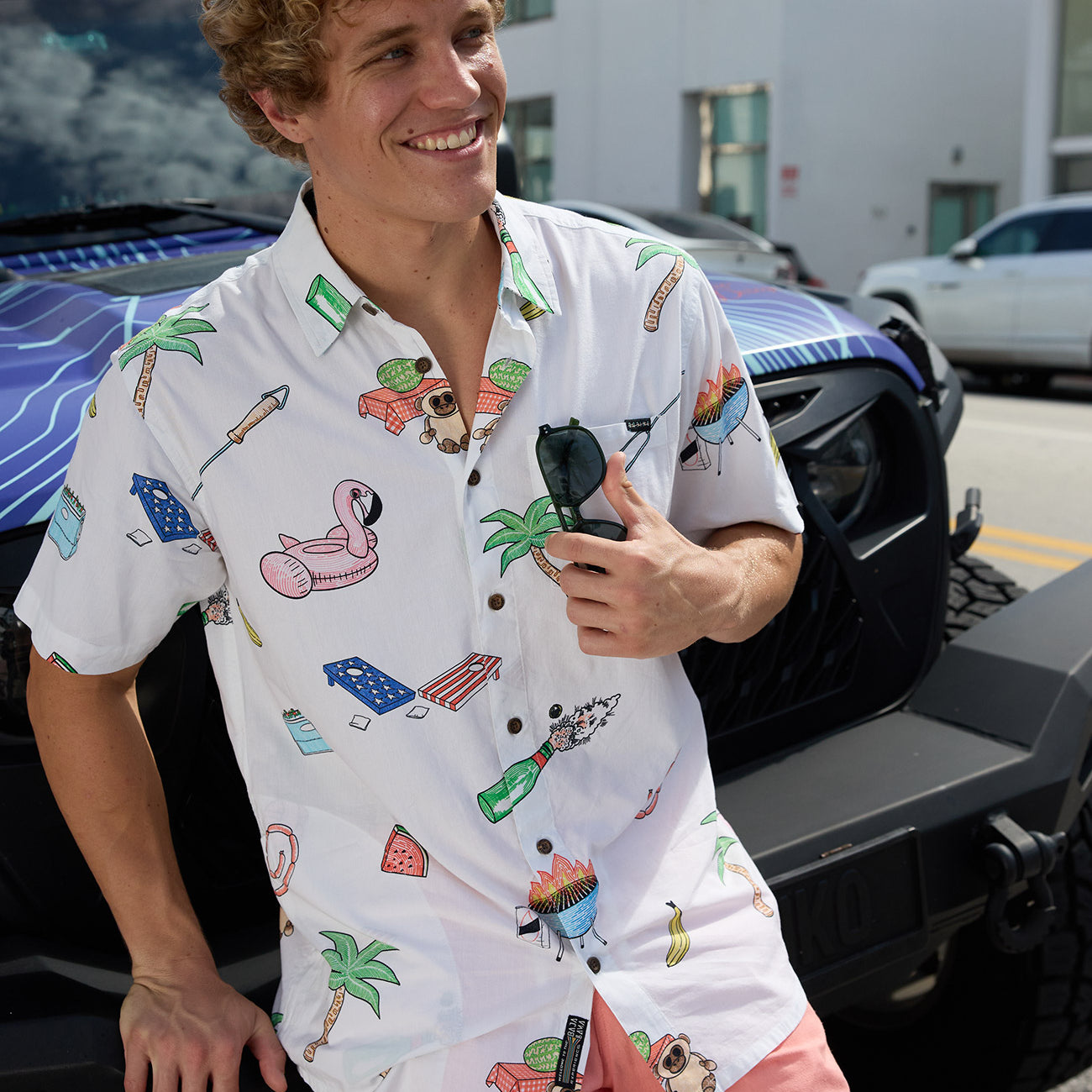 Smiling model leaning against a vehicle wearing the Baja Llama The BBQ Shirt white relaxed-fit button-up with playful cookout and palm tree prints