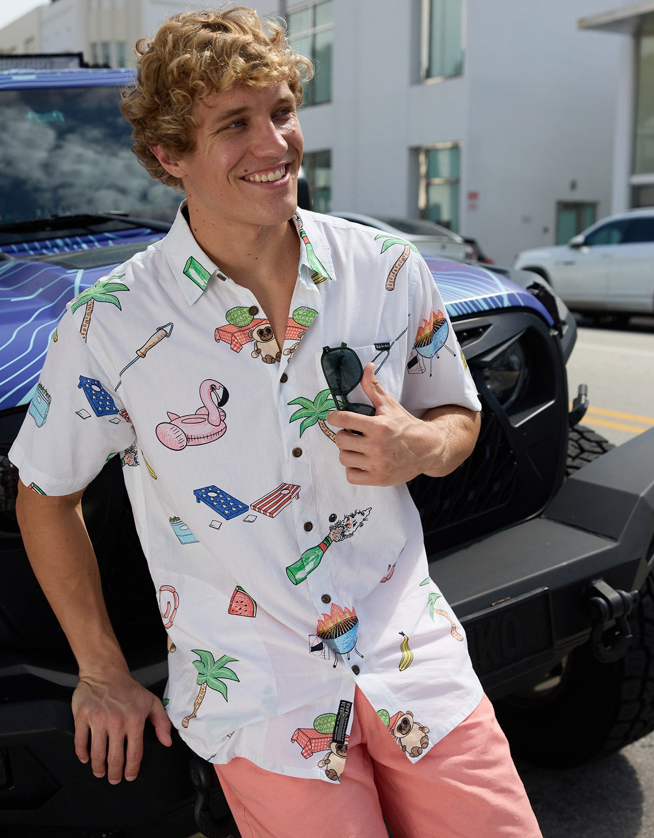 Smiling model leaning against a vehicle wearing the Baja Llama The BBQ Shirt white relaxed-fit button-up with playful cookout and palm tree prints