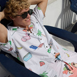 Relaxed male model wearing sunglasses and the Baja Llama The BBQ Shirt white button-up with all-over summer cookout graphics while lounging in a chair outdoors