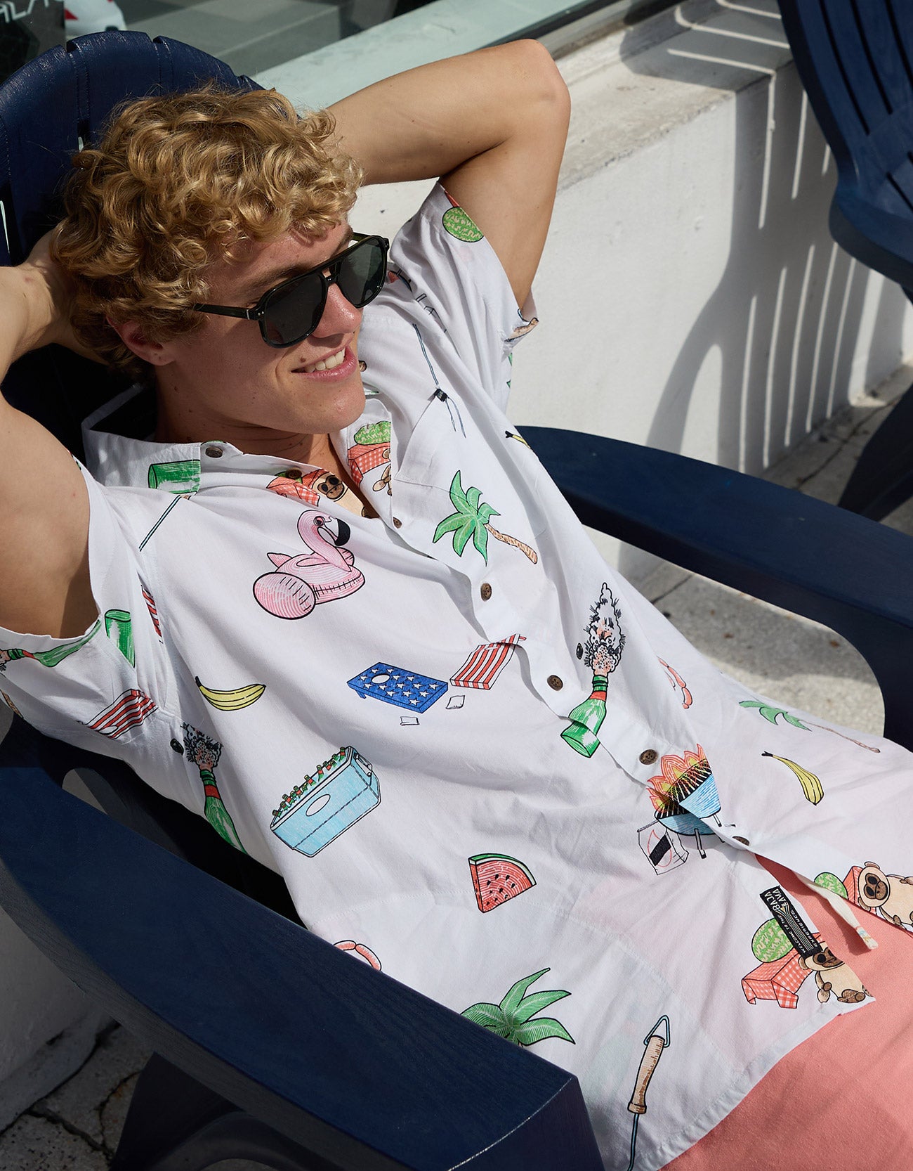 Relaxed male model wearing sunglasses and the Baja Llama The BBQ Shirt white button-up with all-over summer cookout graphics while lounging in a chair outdoors