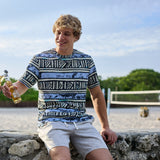Smiling model sitting on a stone wall near a beach volleyball court holding a bottled drink, wearing The Good Ol’ Days patterned heavy tee