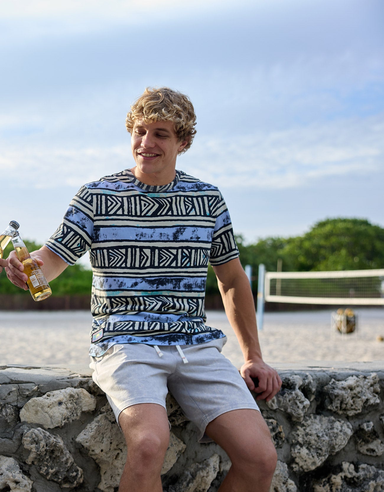 Smiling model sitting on a stone wall near a beach volleyball court holding a bottled drink, wearing The Good Ol’ Days patterned heavy tee