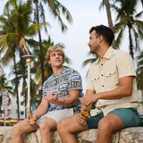 Two male models sitting on a stone wall under palm trees holding bottled drinks, wearing Baja Llama apparel including The Good Ol’ Days patterned heavy tee
