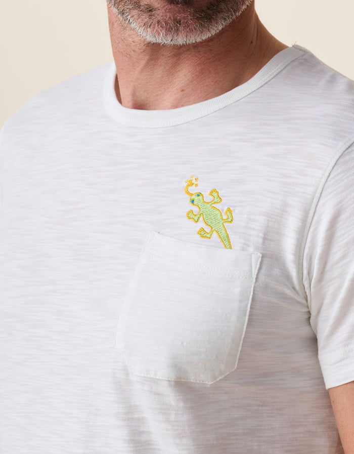 A close-up shot of the left chest on the Baja Llama Thin Lizzy Pima Slub Pocket Tee as worn by a model, showing the textured white fabric and a patch pocket topped with a small embroidered lime green lizard design.