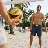 Baja Llama Too Easys Dark Slate Stretch Cotton Shorts lifestyle shot of a man smiling on a sandy beach playing volleyball.