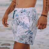 White men's swim trunks featuring tropical palm trees and taro in different hues of blue.