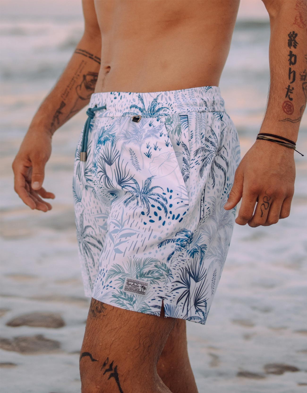 White men's swim trunks featuring tropical palm trees and taro in different hues of blue.